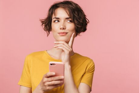 A woman holds her phone and, with her hand on her chin, looks to her side, smiling subtly and thinking.